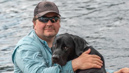 Photo of Tim and his guide dog Harlow, sitting in a boat with the water in the background.