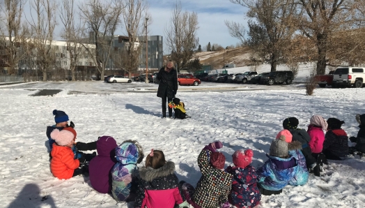 Un groupe d'enfants assis dans la neige devant un éleveur de chiots bénévole et un Labrador Retriever noir portant un nœud papillon de clown et un gilet jaune de futur chien-guide.