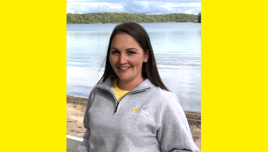 Lindsay Garrett, Program Manager, is standing on the Lake Joe beach modelling a gray quarter zip sweatshirt.
