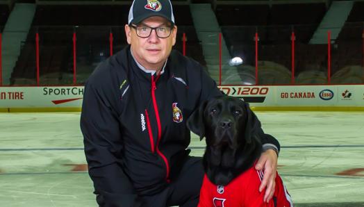 Mark Kelly kneeling next to Rookie, a Future CNIB Guide Dog wearing an Ottawa Senators jersey, with his arm around Rookie while they both smile for the camera. They are on a red carpet laid over the ice of the Canadian Tire Centre, home to the Ottawa Senators.