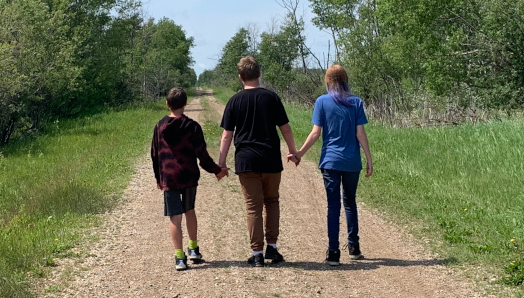 The backs of Ashley’s three children as they walk down a secluded road in a lush, green area. The children are holding hands.