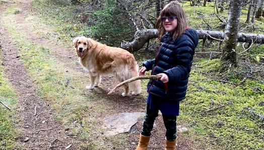 Rhea and her CNIB Buddy Dog Ivy, a two-year-old golden retriever, walking along a trail in the woods. Rhea is holding a stick and they are both looking back at the camera, smiling.