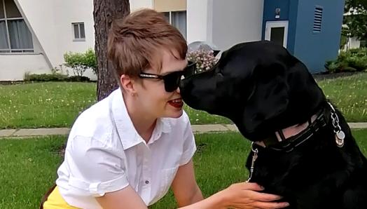 Denise, assise dans l'herbe, vêtue d'une jupe jaune vif, nez-à-nez avec Tara, son chien-guide d’INCA. 