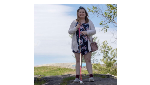 Alicia Chenier sourit et pose pour une photo en plein air. Il y a des arbres verts luxuriants et un ciel bleu en arrière-plan. Elle tient sa canne blanche dans sa main droite.