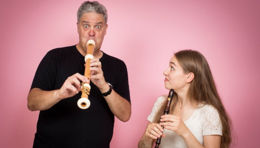 A man and a woman are playing recorders. The man has a funny look on his face, and the woman is smiling