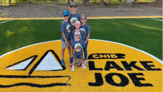Cinq enfants posent sur le logo jaune géant d’INCA au milieu du nouveau terrain de soccer du camp Lake Joe. Ils portent tous un t-shirt gris à l’effigie du camp Lake Joe d’INCA