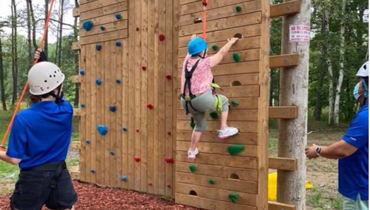 Fran Moreau in her helmet and climbing harness scaling the climbing tower while CNIB Lake Joe staff assist.