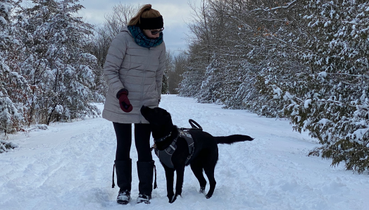 Kathleen, wearing a parka while walking down a snowy trail in the forest with her CNIB Guide Dog, Lily