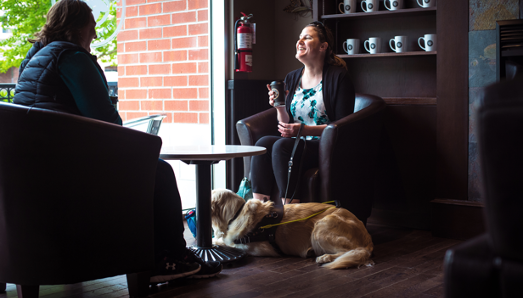 Larissa est assise sur une chaise en face d’une autre personne dans un café, près de la fenêtre avec son chien-guide Piper couché à ses pieds.
