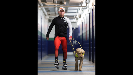Kevin portant des patins à glace et des vêtements de sport marchant dans un couloir avec son chien-guide.