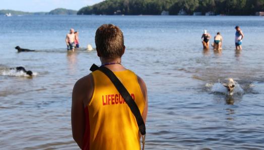 Sauveteur face à l’eau où jouent des personnes accompagnées de chiens-guides. Il porte un maillot jaune avec le mot « Sauveteur » en anglais en rouge.