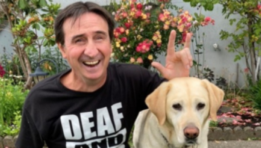 Eddy sits in front of a backyard garden with his guide dog. His left-hand hovers above the dog’s head as he signs “I love you” in ASL. The black t-shirt Eddy is wearing has a bold text design that reads “Deaf and loud!” 