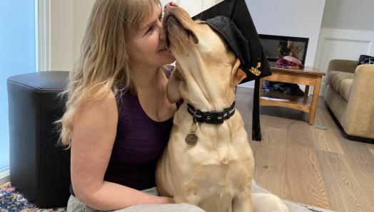 Laurie sits on her living room floor with her guide dog, Bridget, a yellow Labrador retriever-golden retriever cross. Bridget wears a graduation cap and sits in Laurie’s lap. Laurie affectionately presses her face against Bridget’s fluffy face.