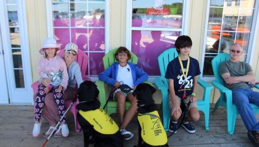 12-year-old Gabriel sits in a colouful Muskoka chair, relaxed and smiling, surrounded by other happy CNIB Lake Joe campers and families, and two CNIB buddy dogs in yellow vests.
