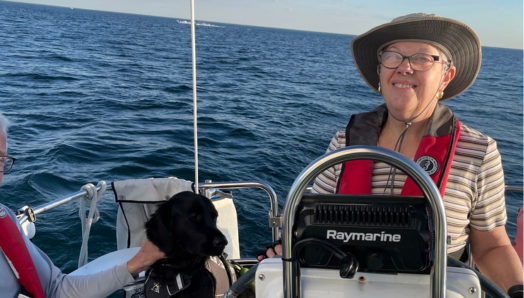 En naviguant sur le lac Ontario, Penny prend la barre du voilier. Elle porte des lunettes, un chapeau et un gilet de sauvetage rouge. Derrière Penny se trouve Honour, son chien-guide noir. À la gauche de Penny, une personne qui est visible en partie porte un gilet de sauvetage. C’est une journée ensoleillée, avec un ciel bleu clair, et le lac est calme.